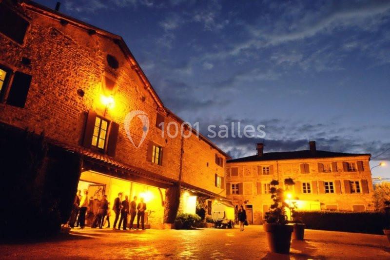 Bâtiments en pierre éclairés par des lumières chaudes au crépuscule, avec des personnes rassemblées à l'entrée.