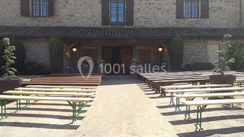 Bancs en bois alignés en extérieur devant une bâtisse en pierre avec des fenêtres et une porte centrale.