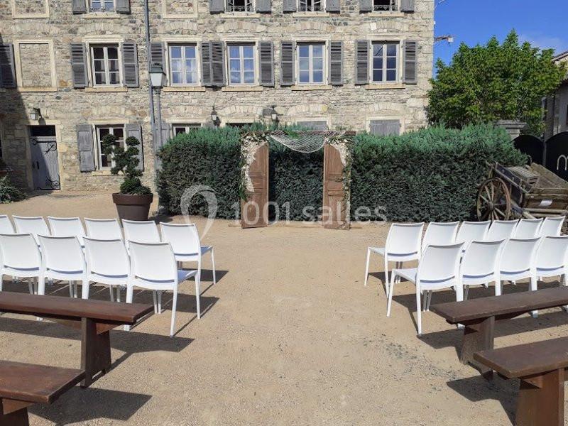Chaises blanches et bancs en bois disposés devant une arche décorée de verdure, dans une cour en plein air.
