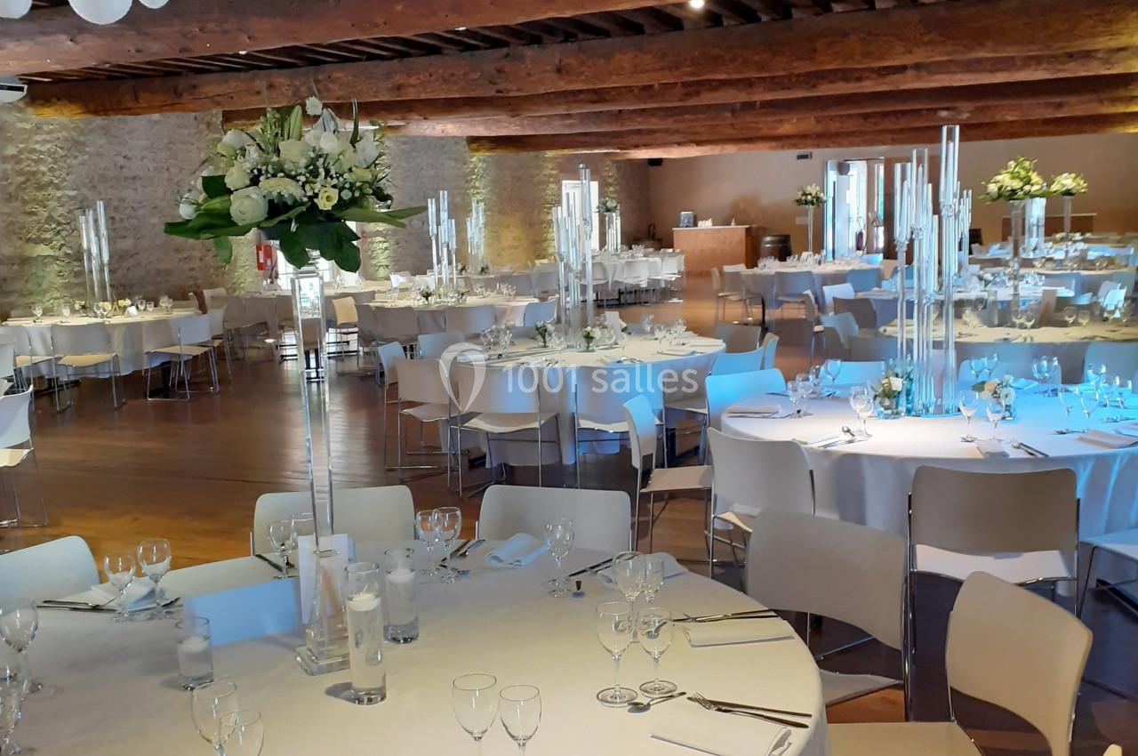 Salle de réception décorée avec des tables rondes dressées, des chandeliers et des fleurs blanches, sous un plafond en bois.