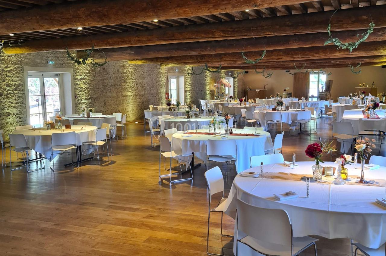 Salle de réception avec tables rondes dressées, nappes blanches, décorations florales et poutres apparentes au plafond.