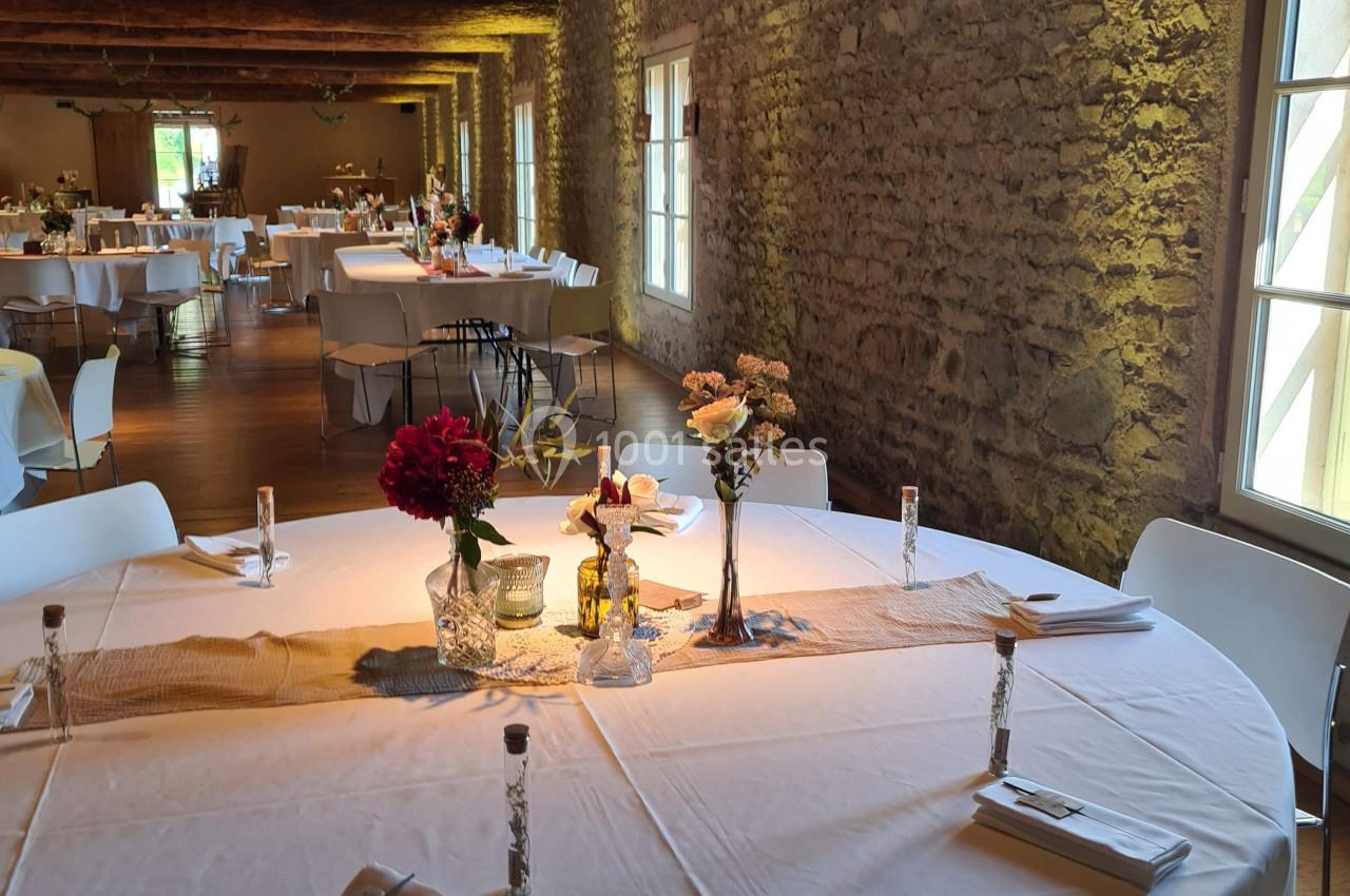 Salle de réception avec murs en pierre, tables rondes dressées avec nappes blanches et décorations florales.