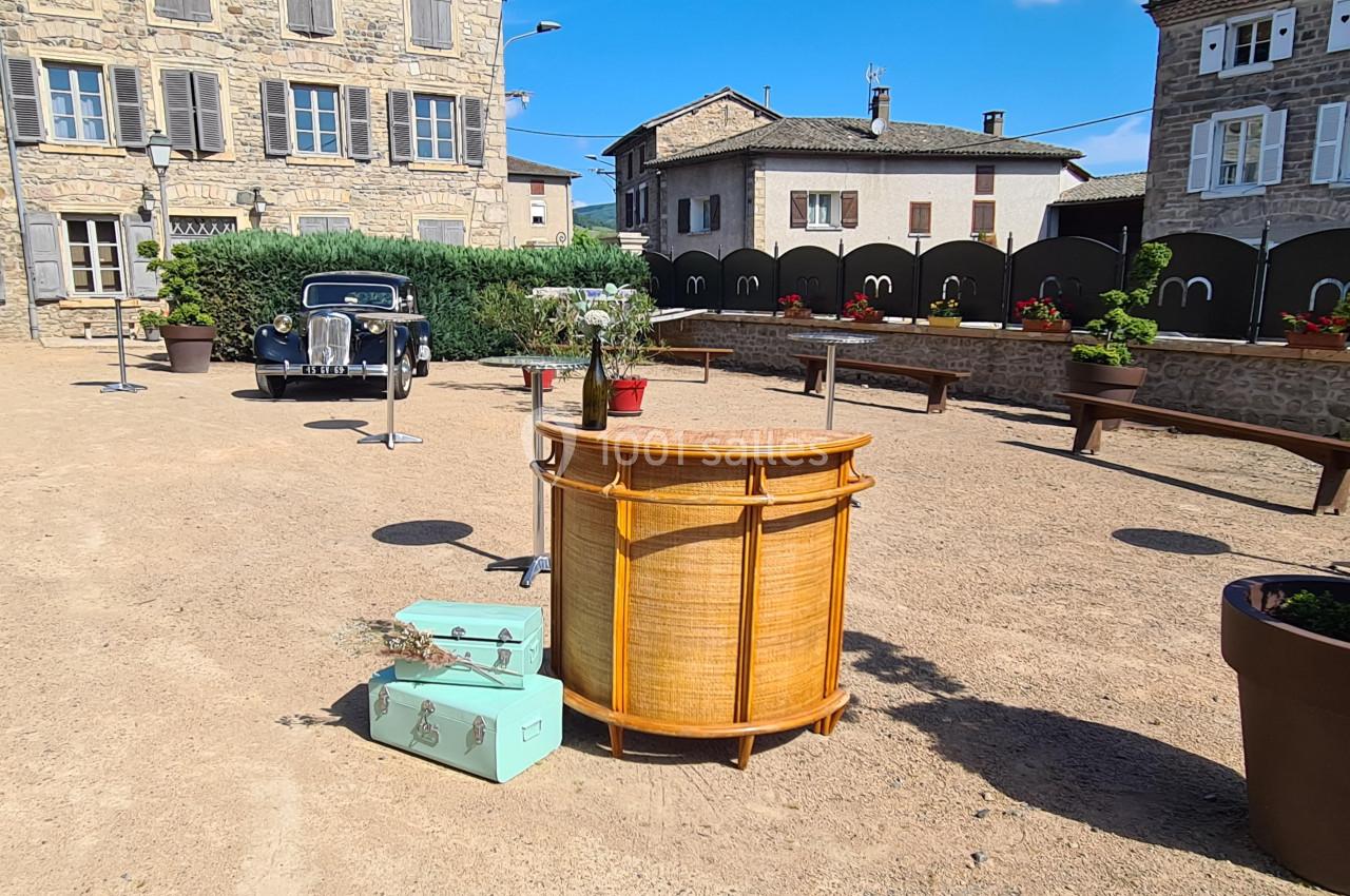 Place ensoleillée avec mobilier vintage, valises turquoise, bar en rotin et voiture ancienne devant des bâtiments en pierre.