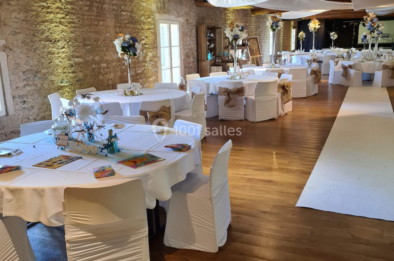 Salle de réception décorée avec des tables rondes, nappes blanches, chaises houssées et arrangements floraux.