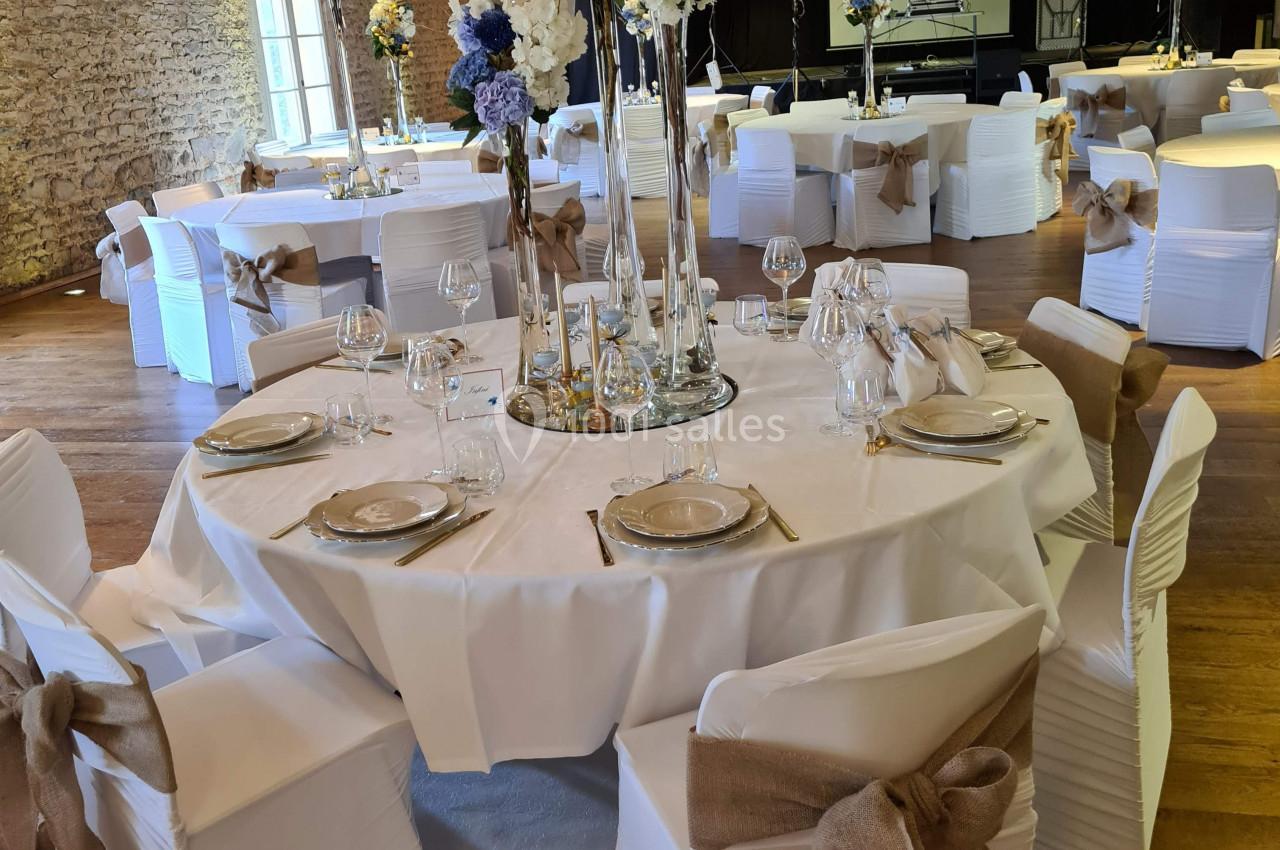 Salle de réception décorée avec des tables rondes, nappes blanches, chaises ornées de nœuds en toile de jute et centres de…