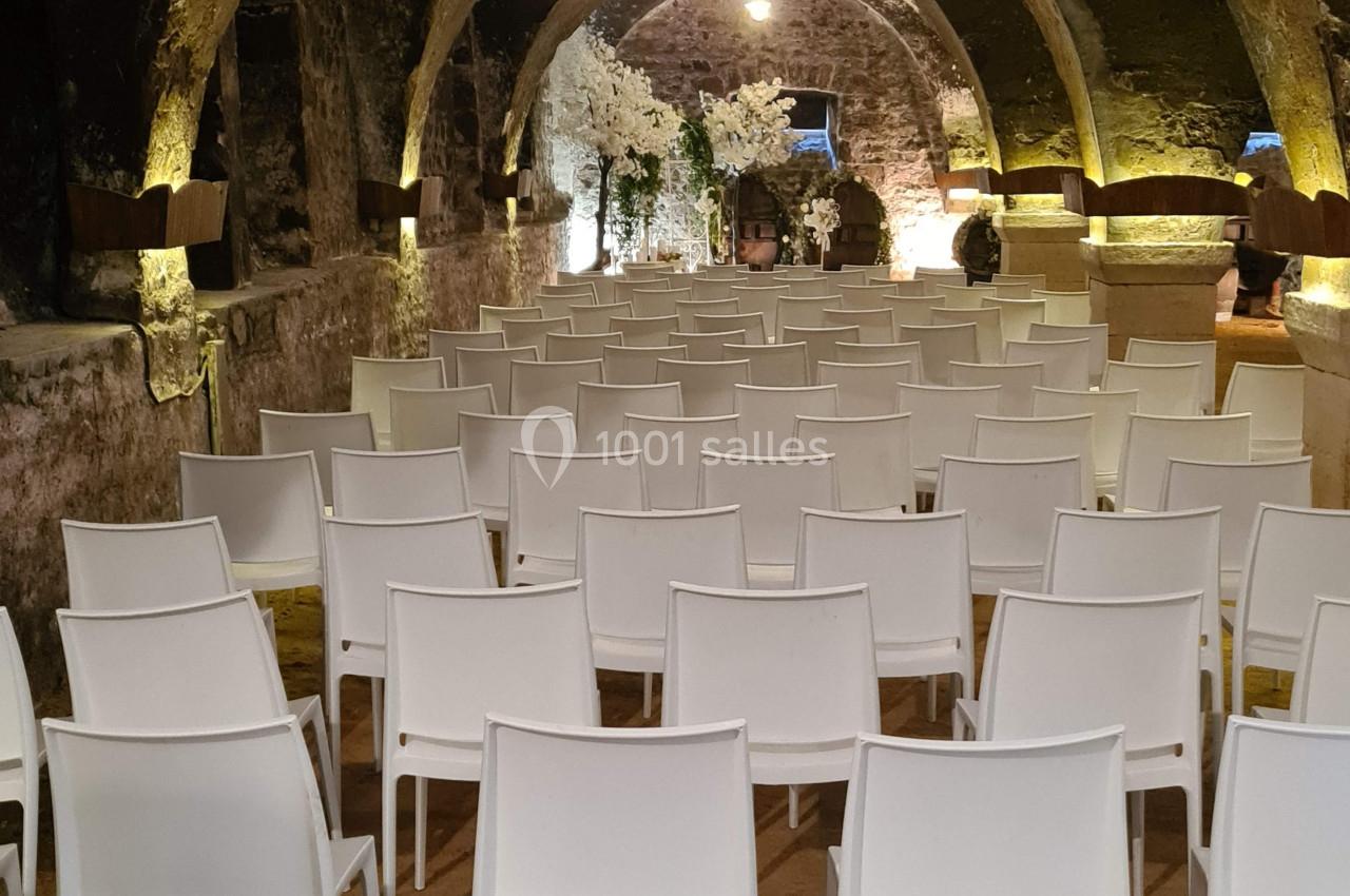 Salle voûtée en pierre avec des rangées de chaises blanches alignées face à une scène éclairée.