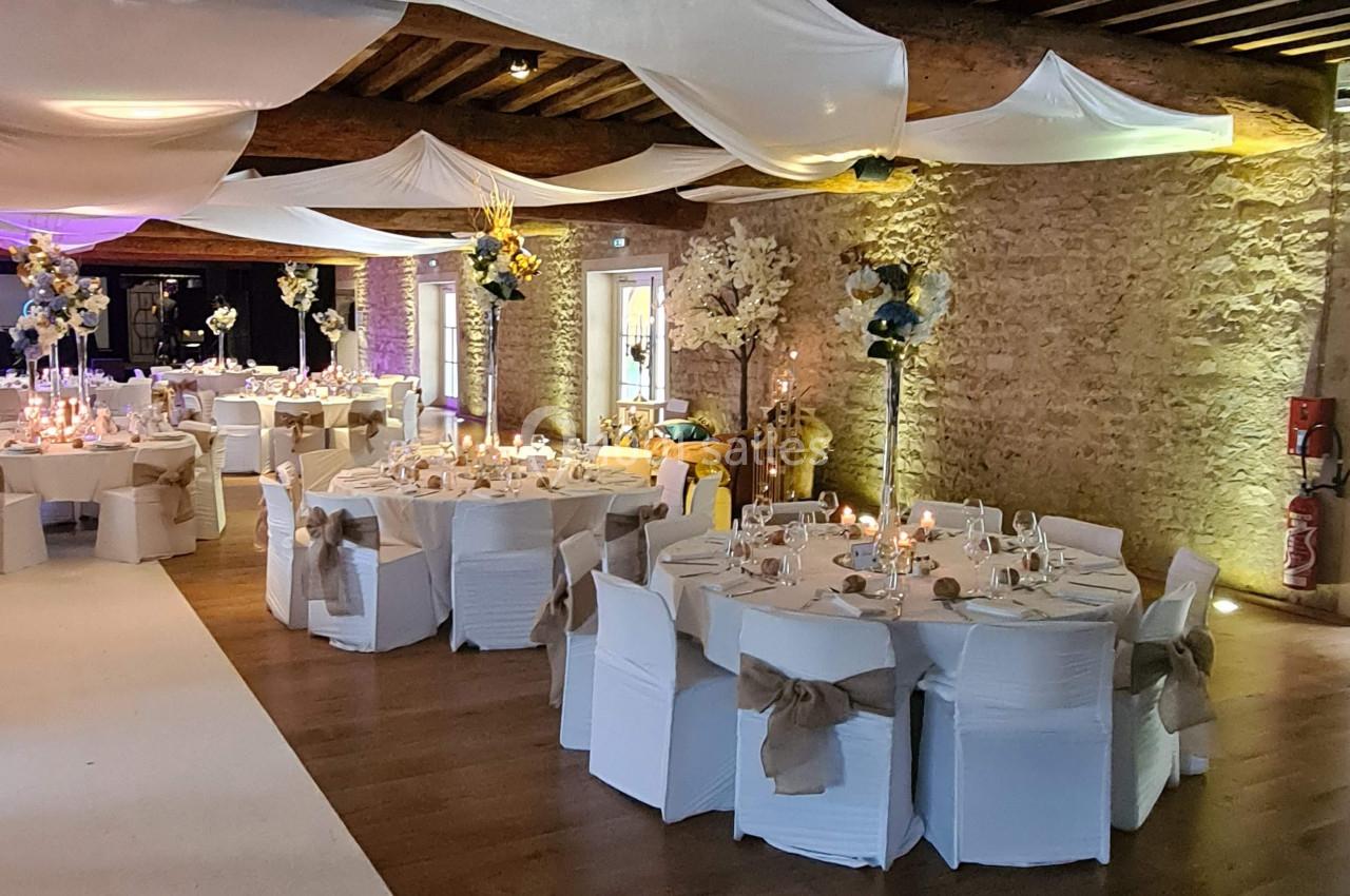 Salle de réception décorée avec des tables rondes, nappes blanches, chaises ornées de nœuds et plafond drapé.