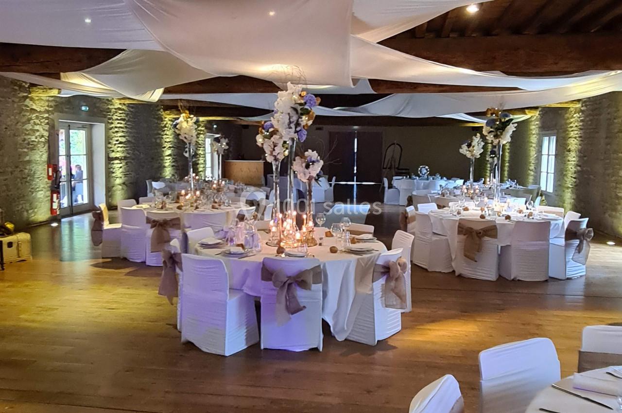 Salle de réception décorée avec des tables rondes, nappes blanches, centres de table fleuris et voilages suspendus.