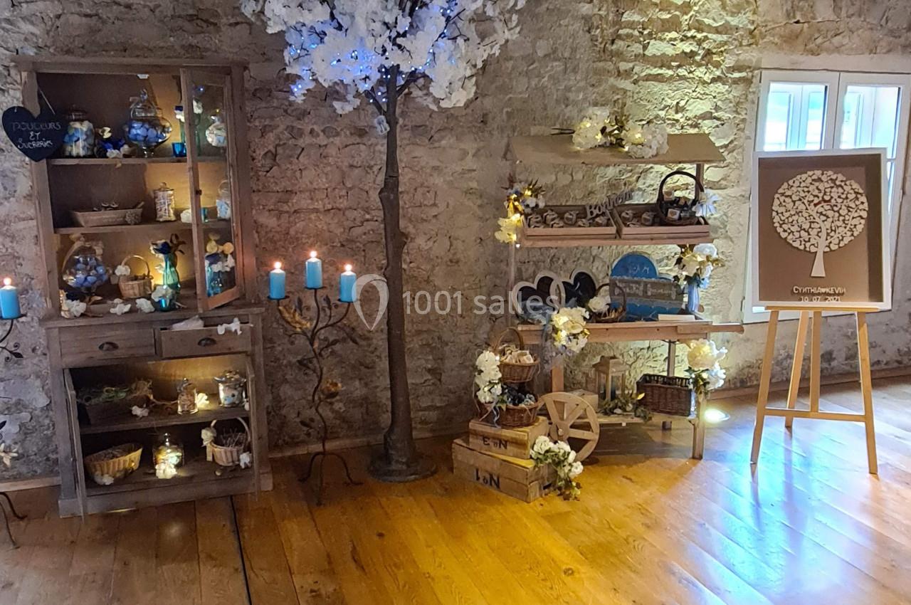 Salle en pierre avec décoration rustique, étagères illuminées, arbre artificiel fleuri et tableau sur chevalet.