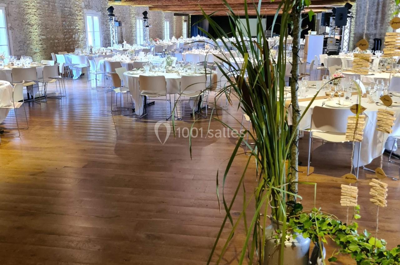 Salle de réception avec tables rondes dressées, poutres apparentes et décoration végétale au premier plan.