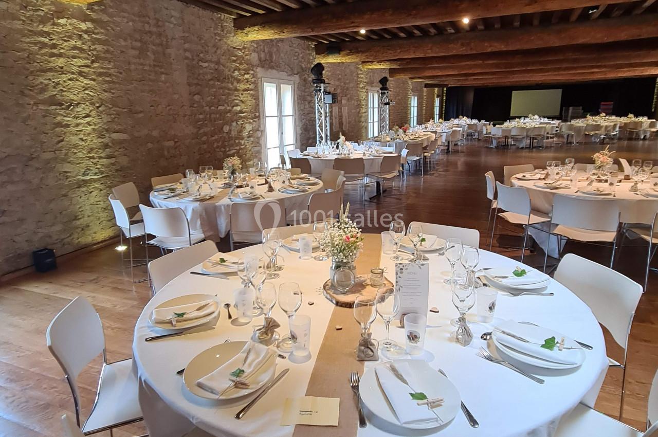 Salle de réception avec tables rondes dressées, nappes blanches, chaises modernes et murs en pierre apparente.