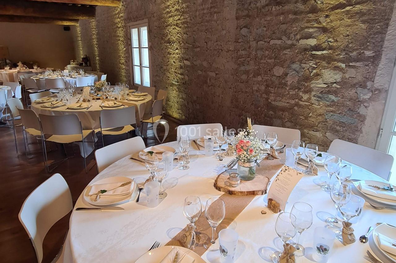 Salle de réception avec tables rondes dressées, nappes blanches, décorations florales et murs en pierre apparente.
