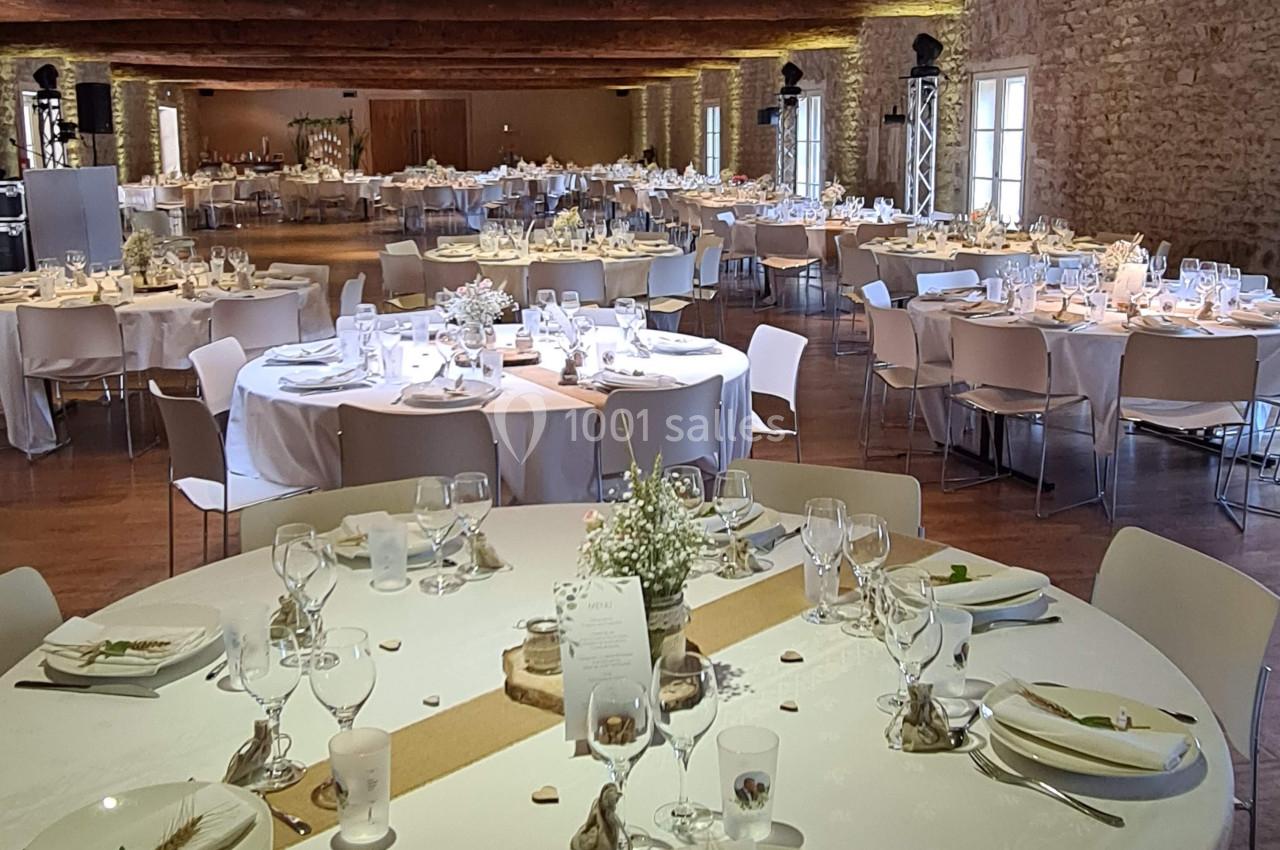 Salle de réception avec tables rondes décorées pour un événement, dans un espace aux murs en pierre et poutres apparentes.