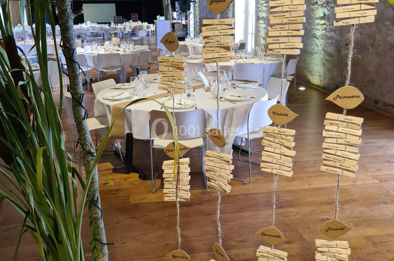 Décoration de mariage avec des marque-places suspendus en bois, tables dressées en arrière-plan dans une salle rustique.