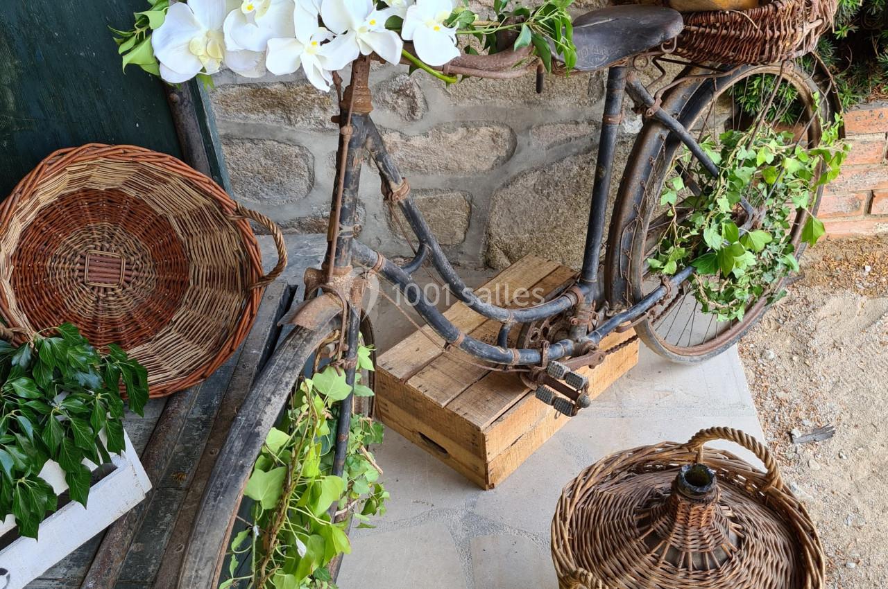 Vélo ancien décoré de fleurs blanches et de feuillage, entouré de paniers en osier sur une terrasse en pierre.