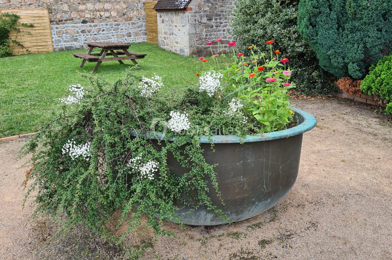 Bac rond en métal contenant des plantes fleuries, situé dans une cour avec pelouse, arbres et bâtiments en pierre.
