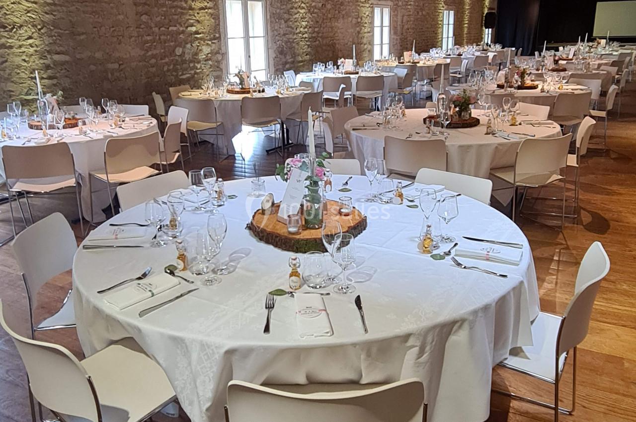 Salle de réception avec tables rondes dressées, nappes blanches, chaises modernes et murs en pierre apparente.