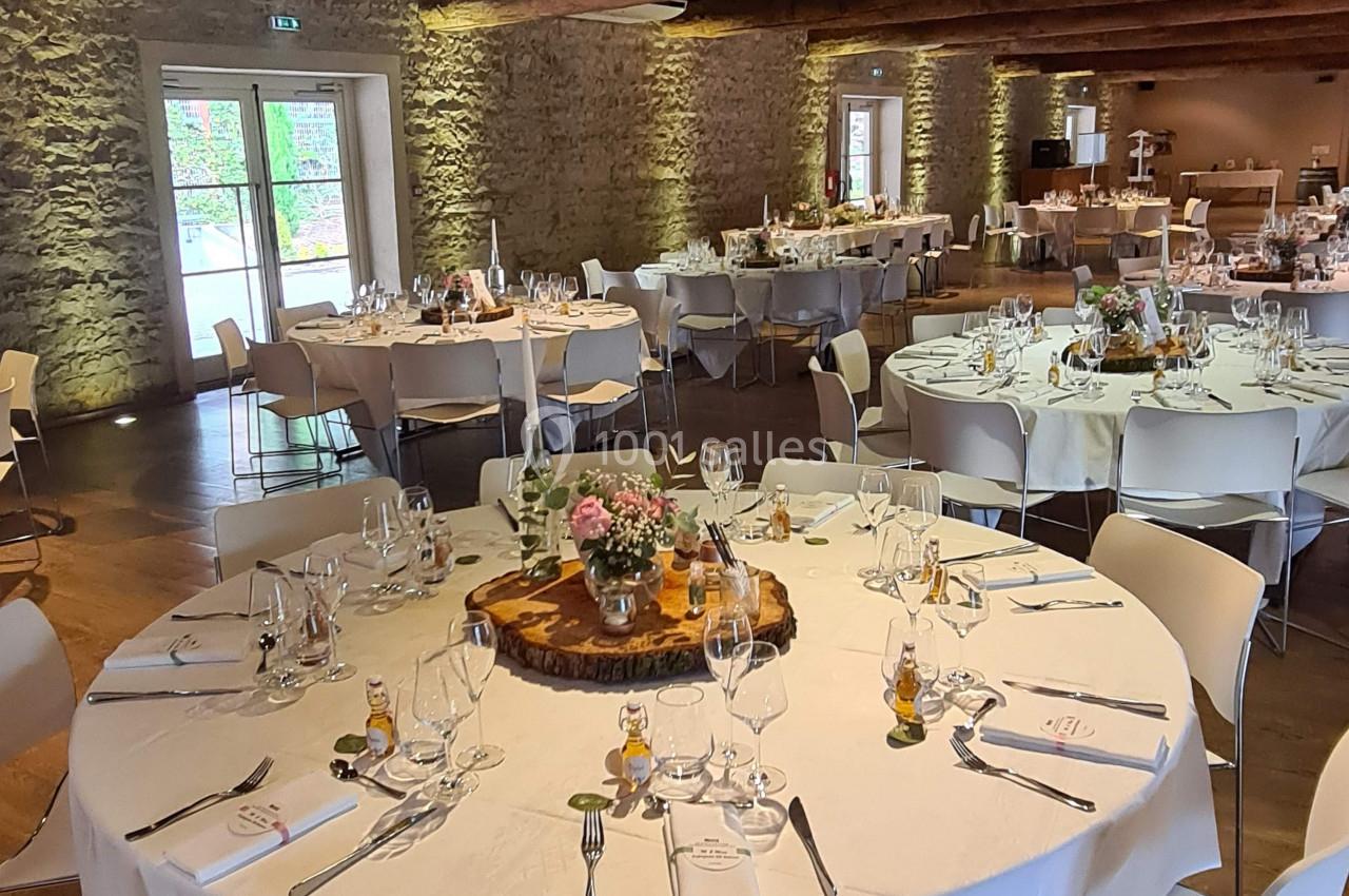 Salle de réception avec tables rondes dressées, nappes blanches, décorations florales et murs en pierre apparente.