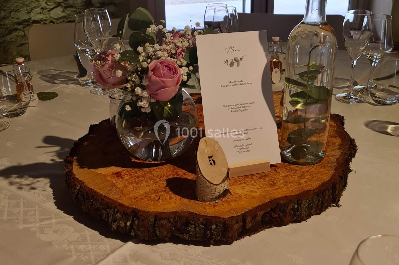 Centre de table avec rondin de bois, vase de fleurs, menu, bougie et bouteille d'eau, sur une nappe blanche.