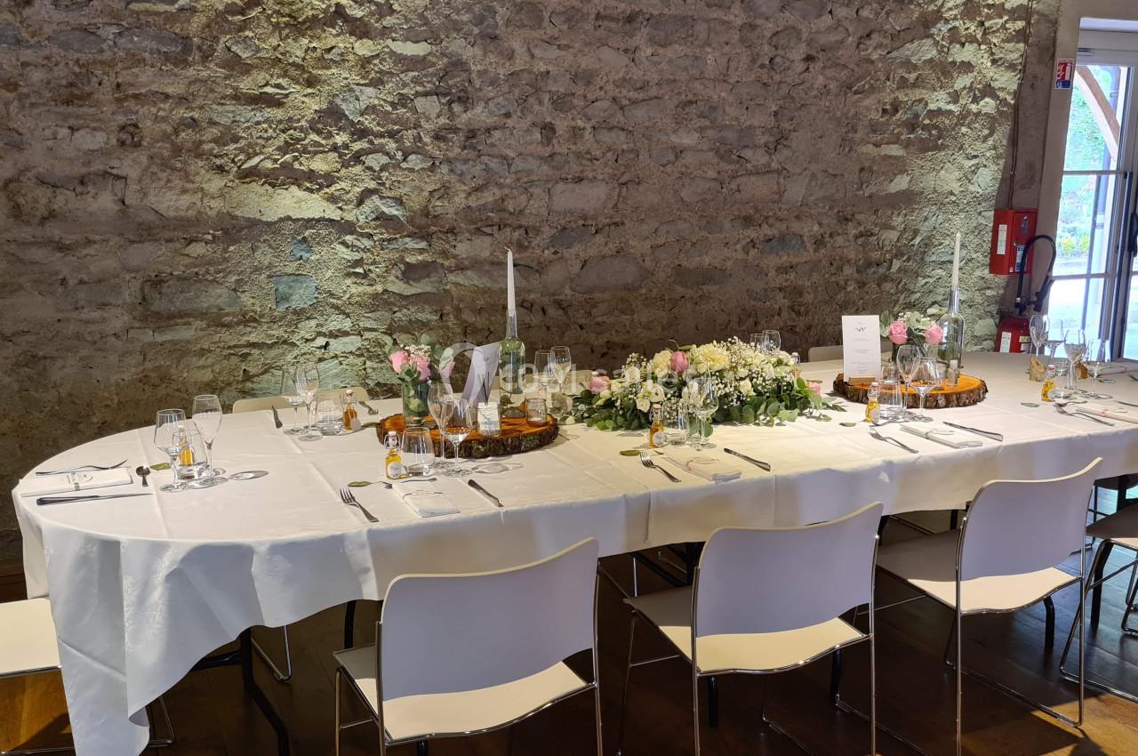 Table élégamment dressée avec nappes blanches, fleurs, bougies et vaisselle, dans une salle aux murs en pierre apparente.