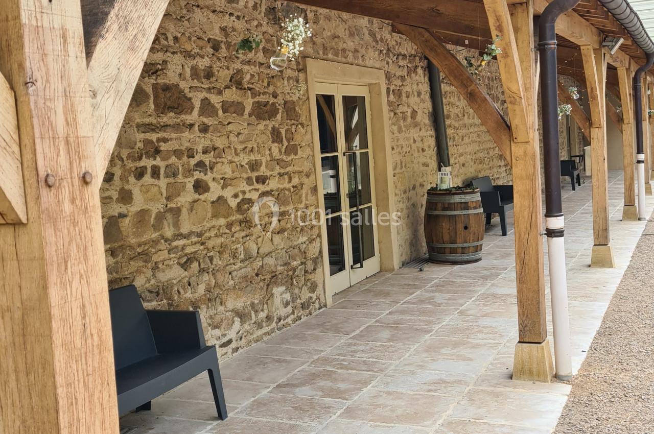 Terrasse couverte avec poutres en bois, murs en pierre, sol carrelé, bancs noirs et tonneau décoratif.