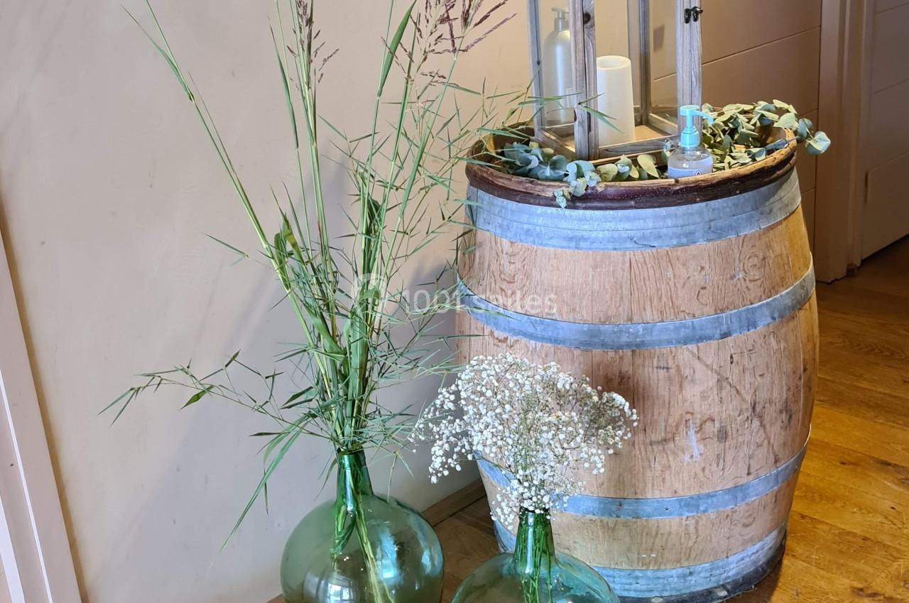 Deux vases en verre vert avec des fleurs séchées posés au sol près d'un tonneau décoré d'une lanterne et de bougies.