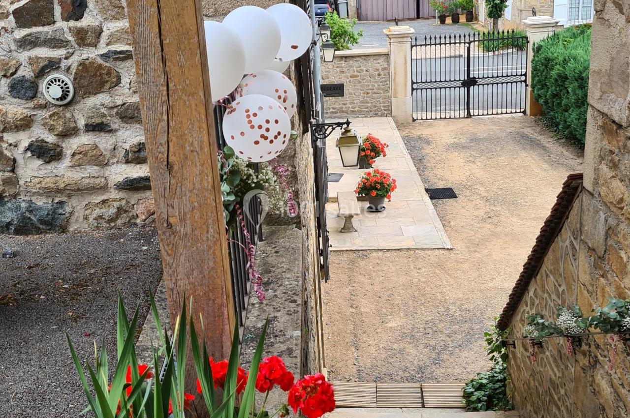 Escalier en pierre bordé de géraniums rouges, menant à une cour avec des ballons blancs et une vue sur des bâtiments en…