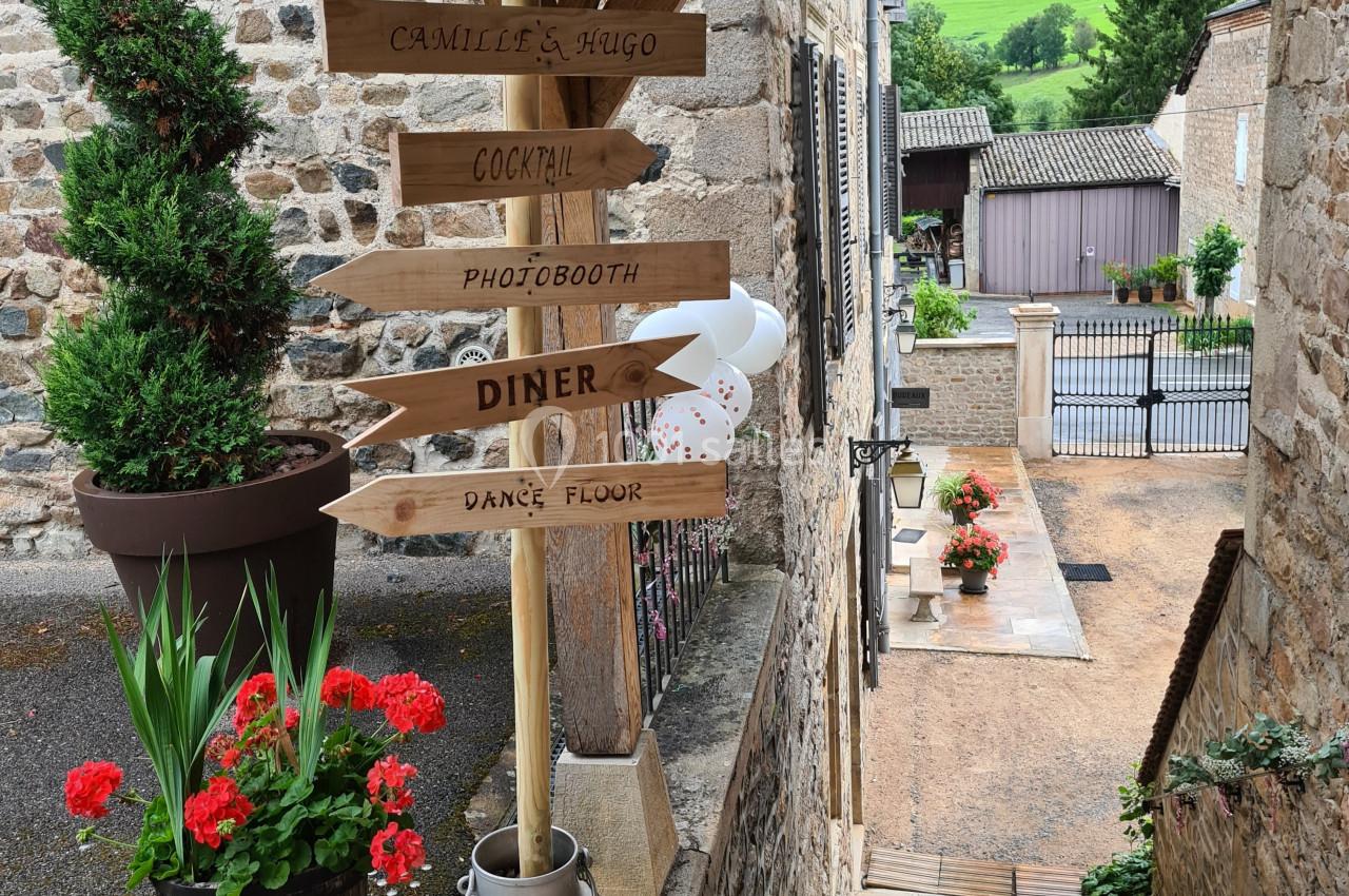 Panneau en bois indiquant des directions pour un événement, dans une ruelle pavée bordée de fleurs et de bâtiments en pierre.