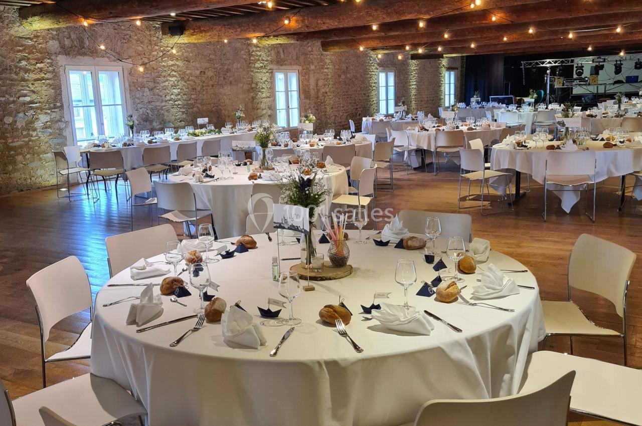 Salle de réception avec tables rondes dressées, nappes blanches, chaises modernes et éclairage chaleureux.