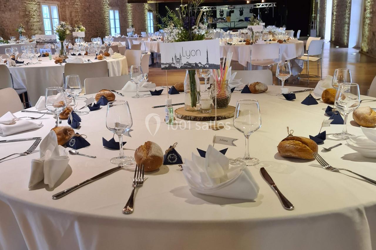 Salle de réception avec tables rondes dressées, nappes blanches, couverts, verres, pains et décorations florales.
