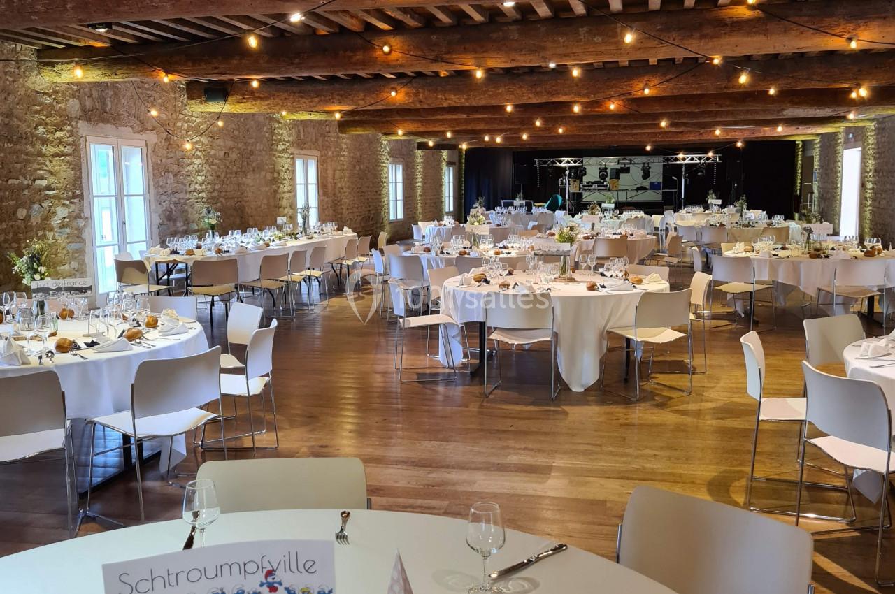 Salle de réception décorée avec des tables rondes dressées, éclairée par des guirlandes lumineuses et des fenêtres en pierre.