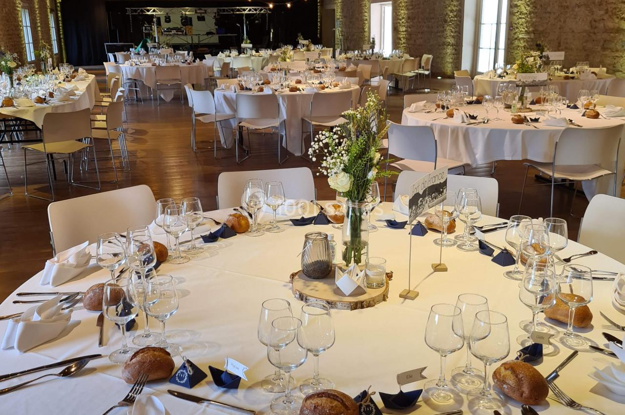 Salle de réception décorée avec des tables rondes dressées, nappes blanches, vaisselle élégante et bouquets de fleurs.