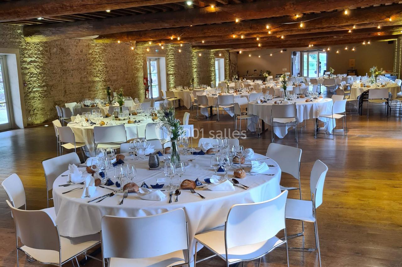 Salle de réception avec tables rondes dressées, nappes blanches et chaises modernes, éclairée par des lumières chaleureuses.