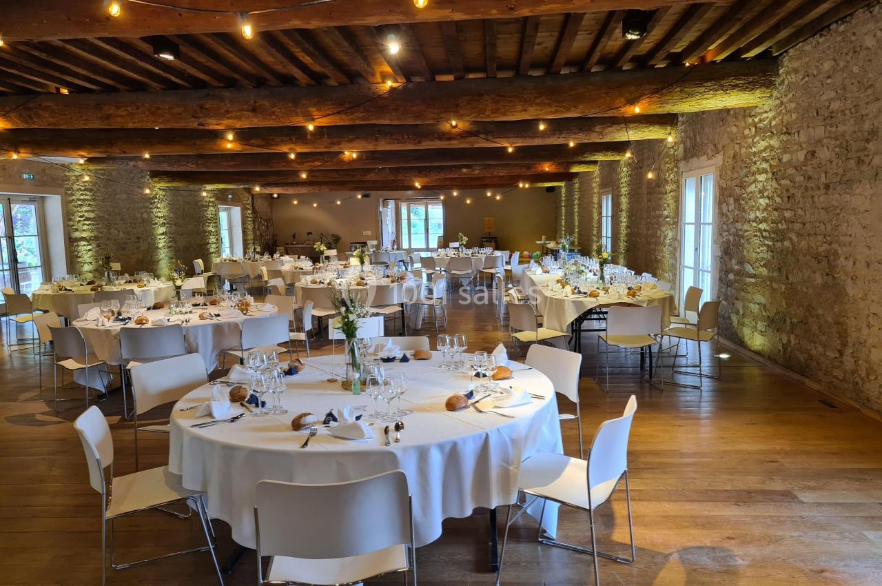 Salle de réception lumineuse avec tables rondes dressées, chaises blanches et décorations élégantes sous un plafond en bois.