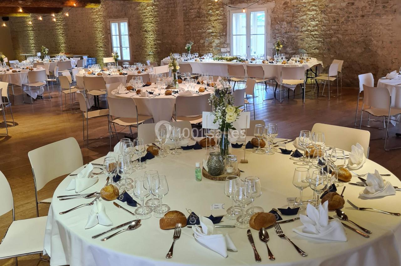 Salle de réception avec tables rondes dressées, nappes blanches, décorations florales et lumière tamisée.