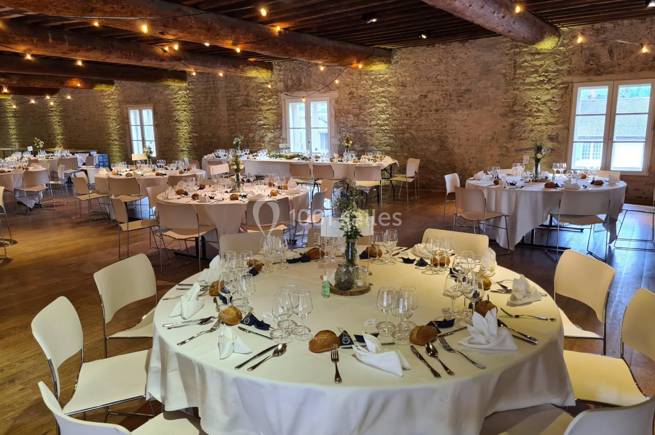 Salle de réception en pierre avec tables rondes dressées pour un repas, décorée de fleurs et éclairée par des guirlandes…