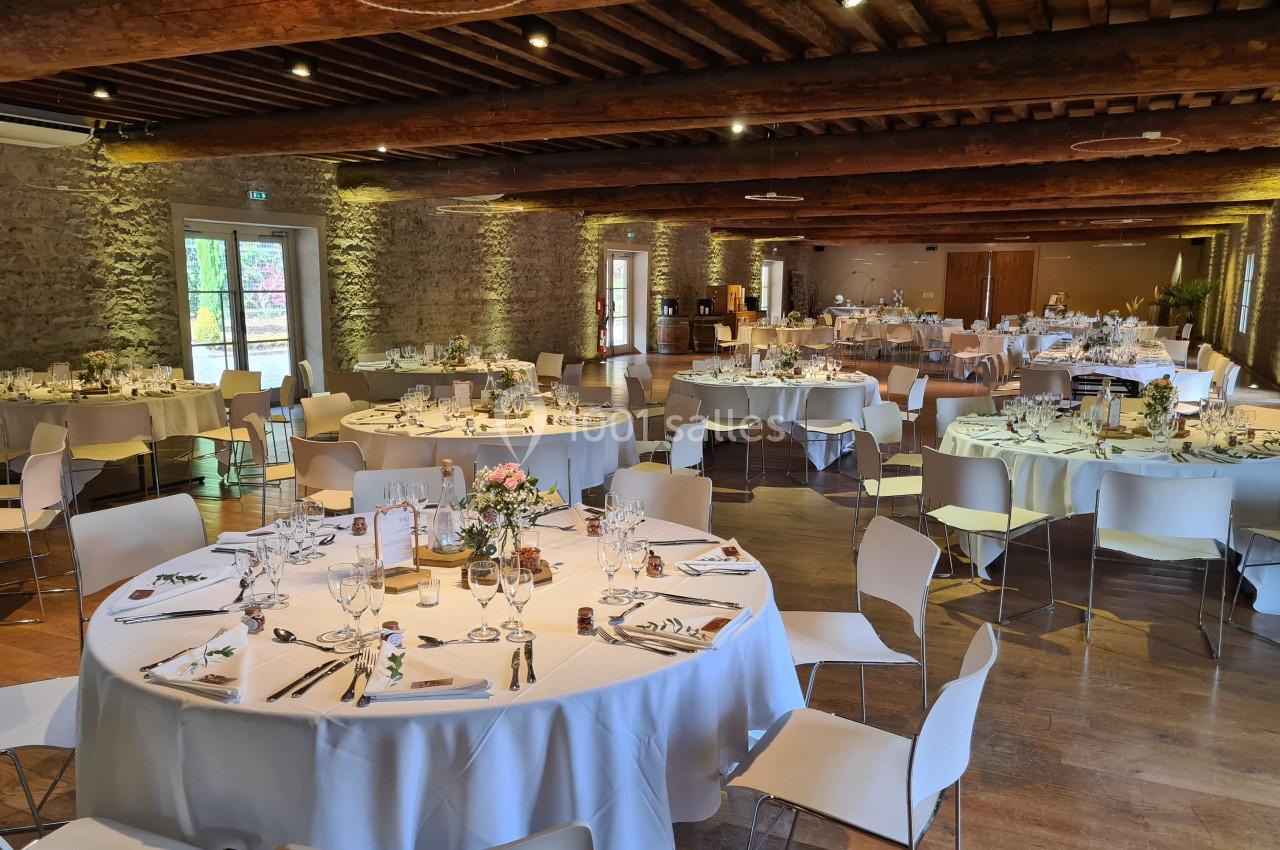 Salle de réception décorée avec des tables rondes dressées pour un événement, dans un cadre en pierre et bois.