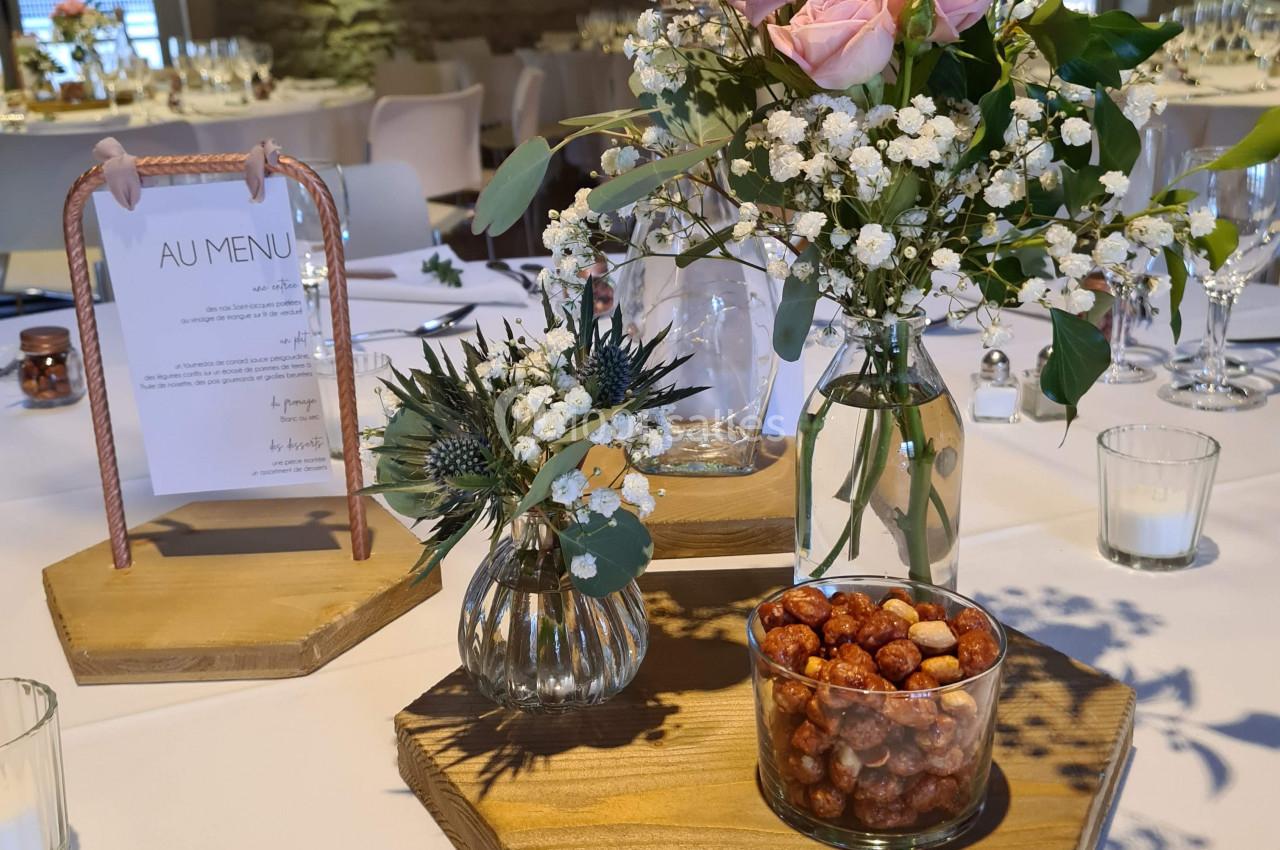 Centre de table avec fleurs roses, gypsophile, pots de fruits secs et menu dans une salle en pierre lumineuse.