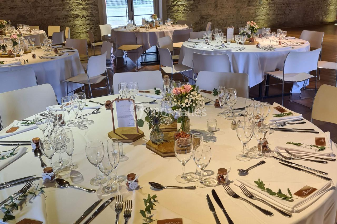 Salle de réception avec tables rondes décorées pour un événement, nappes blanches et arrangements floraux.