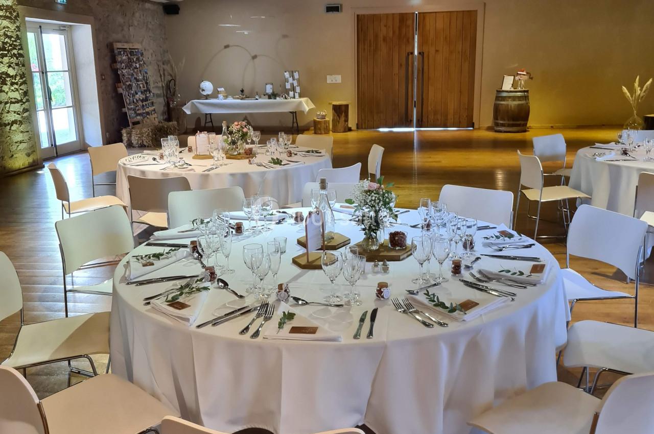 Salle de réception avec tables rondes dressées, nappes blanches, chaises modernes et décoration rustique en bois.