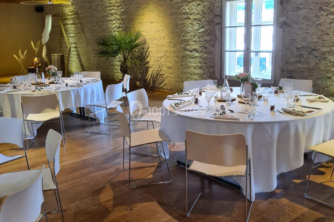Salle de réception avec murs en pierre, tables rondes dressées et chaises blanches sur un parquet en bois.
