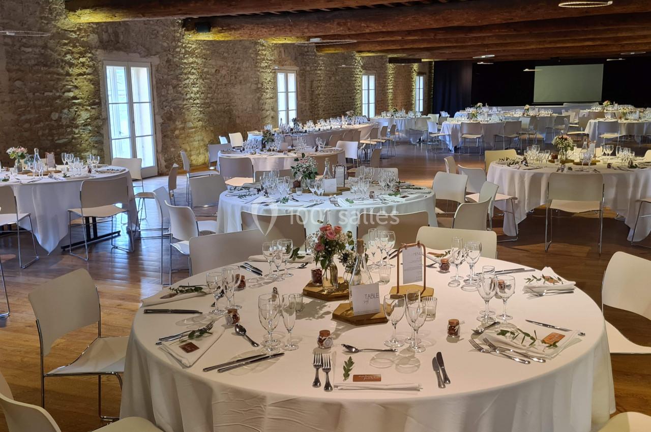 Salle de réception décorée avec des tables rondes dressées pour un événement, éclairée par la lumière naturelle.