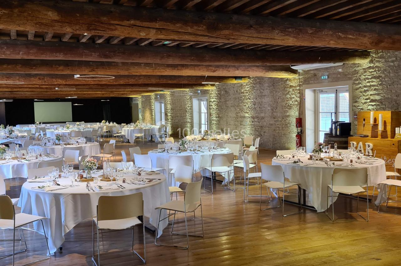 Salle de réception avec tables rondes dressées, chaises blanches, murs en pierre et poutres en bois apparentes.