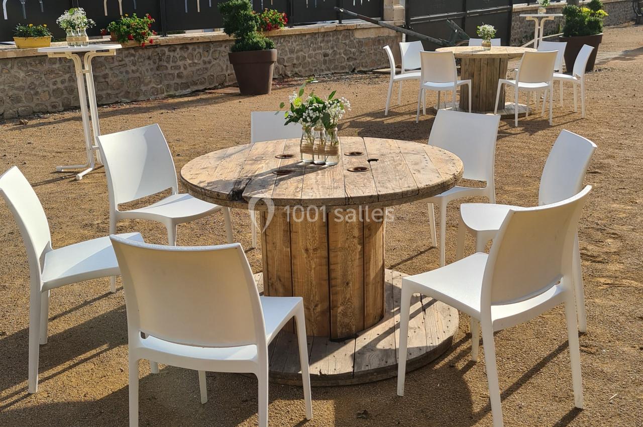 Table ronde en bois de récupération entourée de chaises blanches sur une place extérieure ensoleillée.