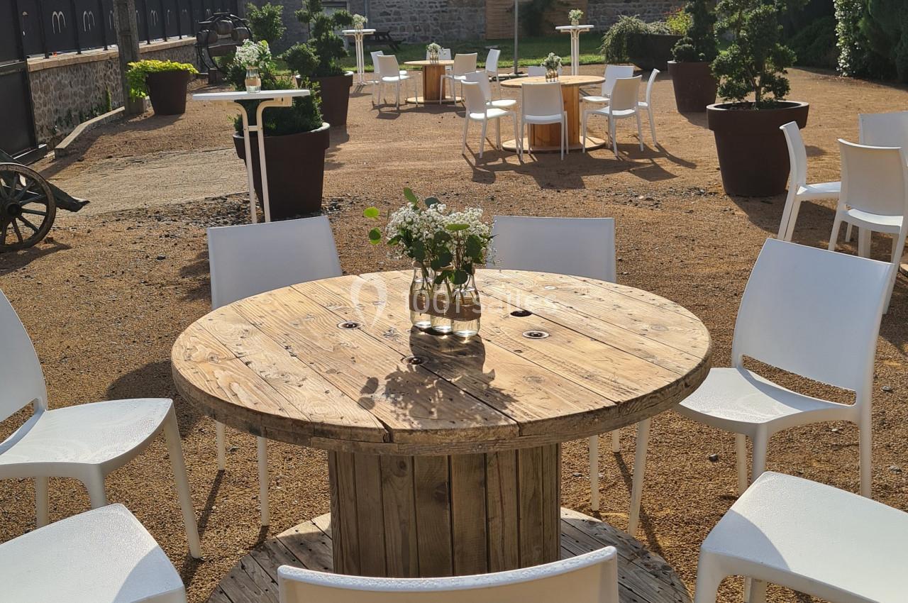 Table ronde en bois entourée de chaises blanches sur une terrasse extérieure en gravier, avec des tables similaires en…
