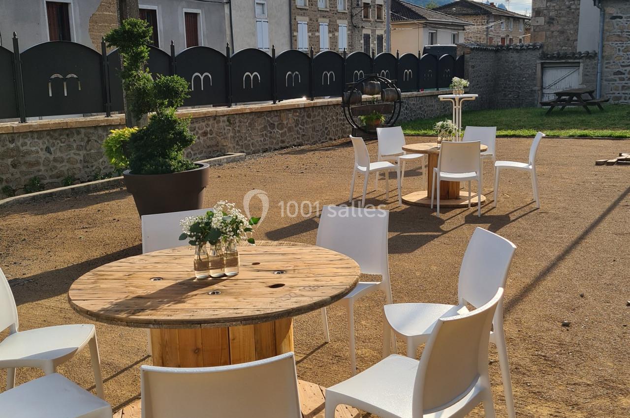 Tables rondes en bois avec chaises blanches disposées en extérieur sur un sol en gravier, entourées de bâtiments.