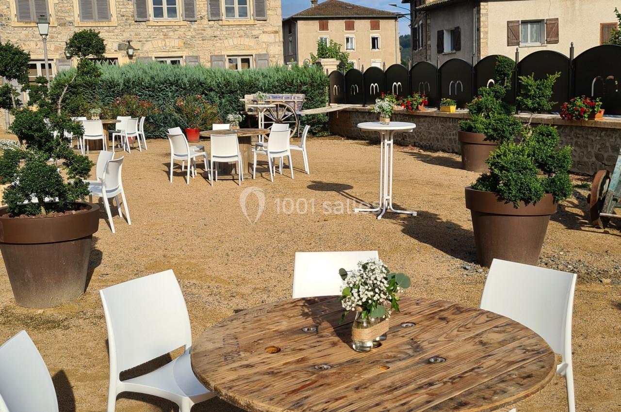 Table ronde en bois entourée de chaises blanches sur une terrasse extérieure avec des bâtiments en pierre en arrière-plan.