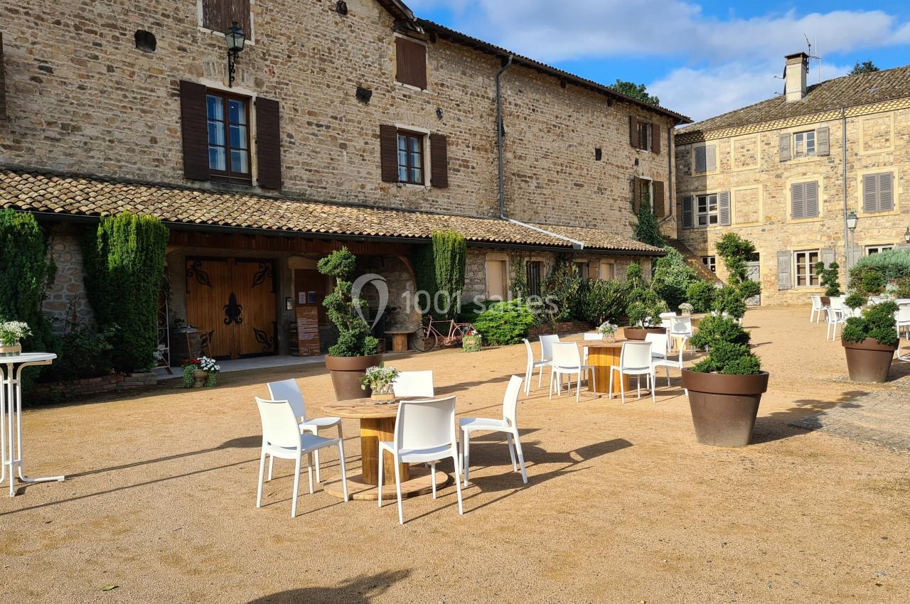 Cour d'une bâtisse en pierre avec des tables et chaises blanches disposées sur un sol en gravier.