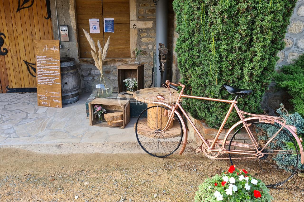 Vélo décoratif en métal cuivré posé devant une entrée en pierre avec plantes, panneaux et caisses en bois.