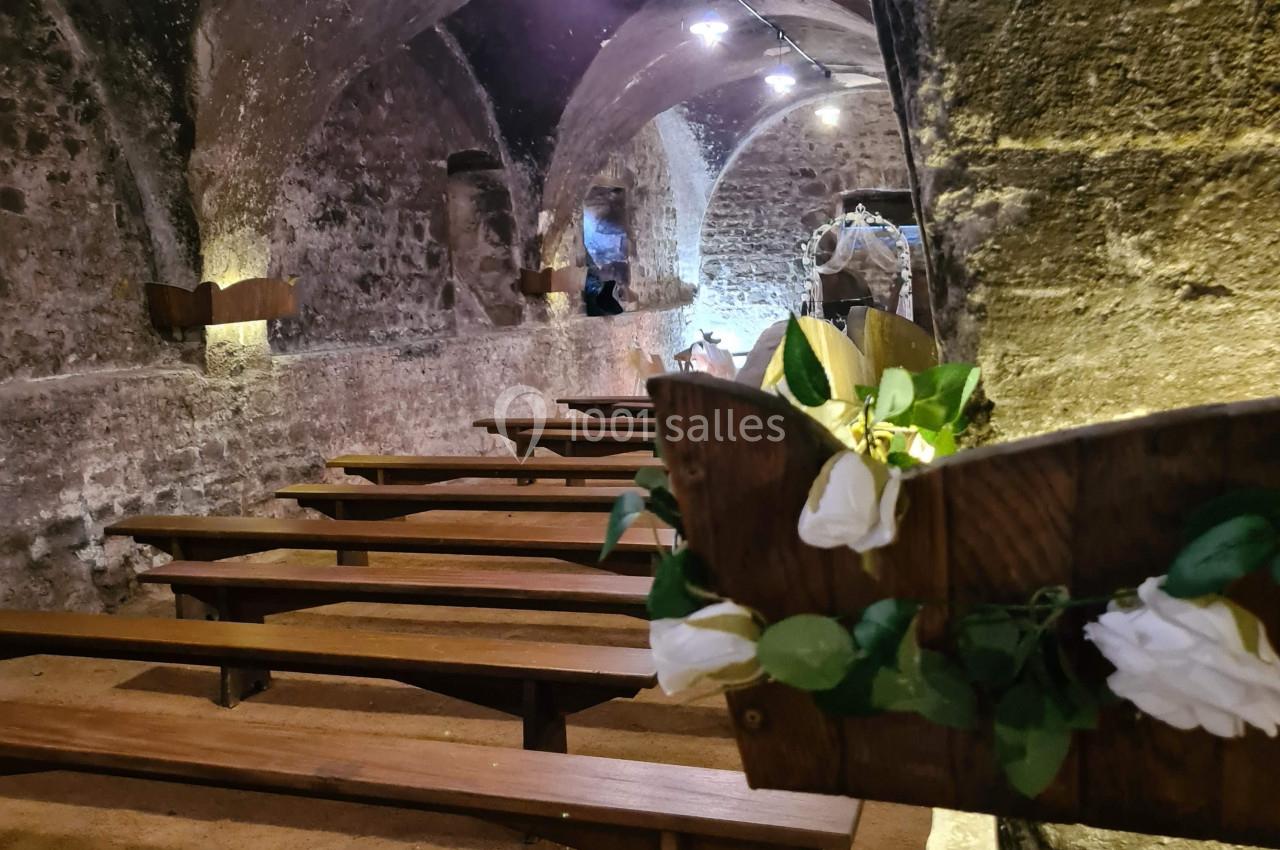 Bancs en bois alignés dans une cave voûtée en pierre, décorée de fleurs blanches et éclairée par des lampes suspendues.